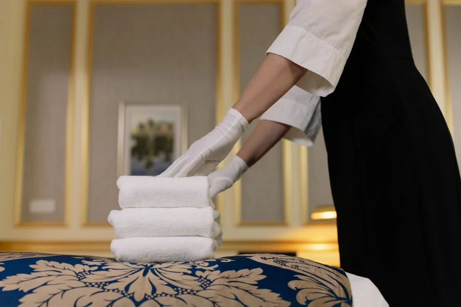 From above, an anonymous housekeeper in yellow rubber gloves sprays cleaner onto a chrome bathroom water faucet